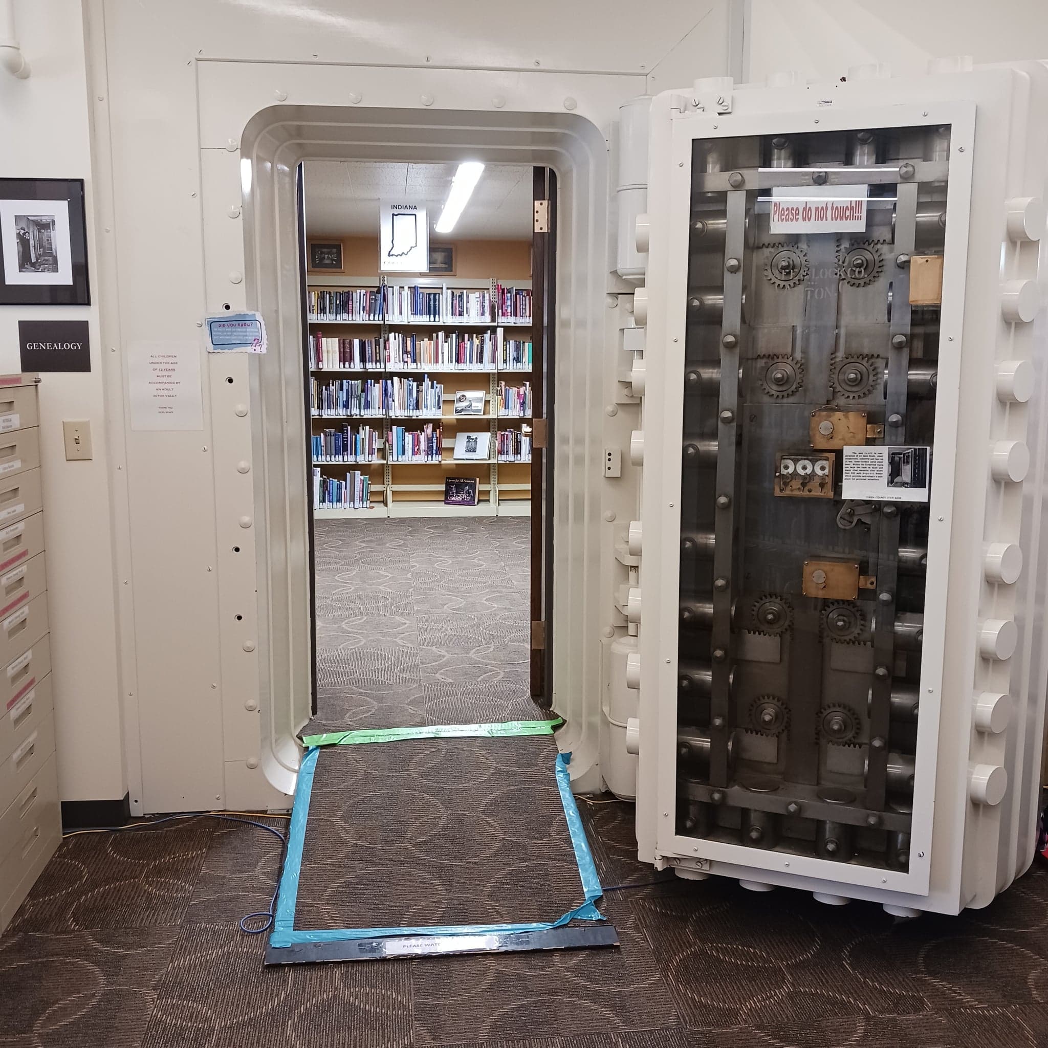 The original bank vault door standing open at the entrance to the genealogy room. A glass panel on its back reveals the brass and steel locking gears, with archive shelves visible through the doorway.